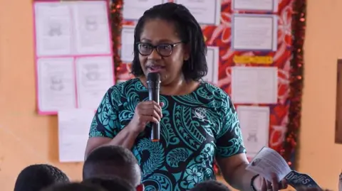 Supplied: Kalesi Volatabu A woman in a black and turquoise dress, wearing glasses and speaking into a microphone in front of several onlookers. Behind her is a wall with several pieces of paper attached to it
