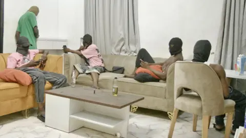 Picture inside the Hustle Kingdom. Four men sit on sofas holding mobile phones. All are wearing balaclavas concealing their faces - two wear pink t-shirts, another a white vest and another is topless. The toom has a marble floor, grey curtains and a coffee table. Another man is standing up looking at a phone with his back to the camera.