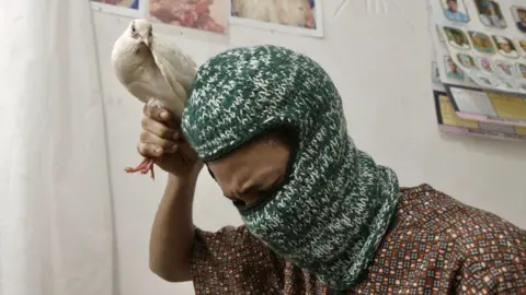 Picture of a man leaning forward and looking down while holding a white dove by its legs, above his head.  The man is wearing a green woollen balaclava and a brown patterned short sleeve shirt.