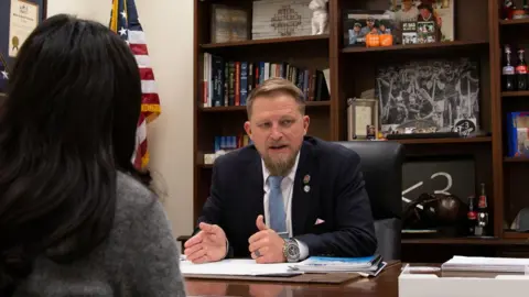 Brandon Guffey pictured sitting behind a desk with bookshelves and a US flag behind him. He has short fair hair and beard, and is wearing a navy suit with a pale blue tie. To the left of the picture is Tir pictured from behind, she has long dark hair and a grey jumper