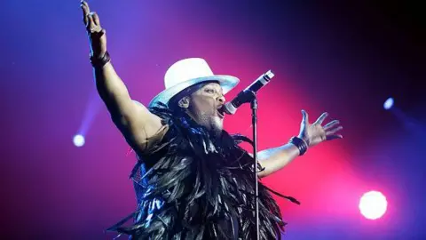 Getty Images D'Angelo, wearing a white hat and a boa with black feathers around his neck, performs on a stage with bright pink lighting. He is raising his arms in the air. 