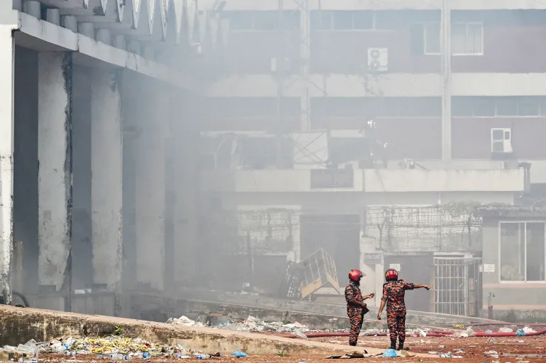 Firefighters inspect as smoke engulfs the fire-damaged cargo terminal of Hazrat Shahjalal International Airport in Dhaka on October 19, 2025, a day after the blaze. A large fire swept through the cargo terminal of Bangladesh's main international airport in Dhaka on October 18, forcing authorities to suspend all flights, officials said. (Photo by Munir UZ ZAMAN / AFP)