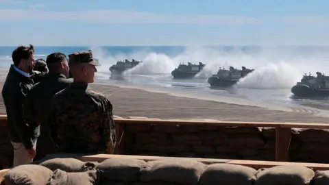 Getty Images JD Vance watches along side military members as boats reach the shores of a beach
