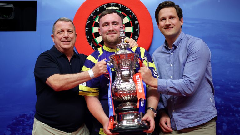 Luke Littler of England poses with The Phil Taylor Trophy alongside Garry Plummer, Owner of Target Darts and James Tattersall, Managing Dire
