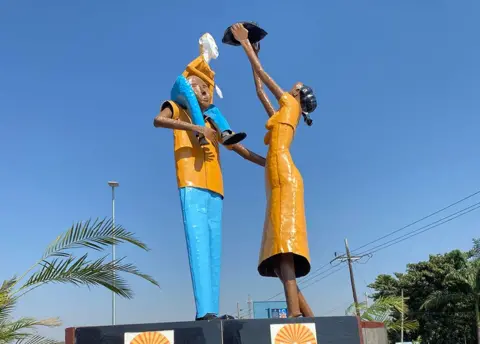A view of a statue showing a woman holding something up to a child who is on a man's shoulders.