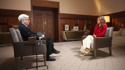 A man with greying hair in a black suit sits to the left interviewing a woman with long brown hair, a red shirt and smart white trousers and shoes