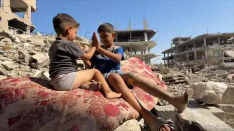 Two young boys, the one on the right wearing a prosthetic leg, clap their hands together while sitting on a carpet on top of the ruins of a building