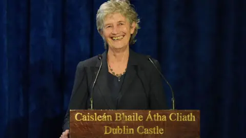 PA Media Catherine Connolly is standing at a podium which says Dublin Castle in both English and Irish. She is wearing a navy suit and silver necklace. She has grey hair and is smiling.