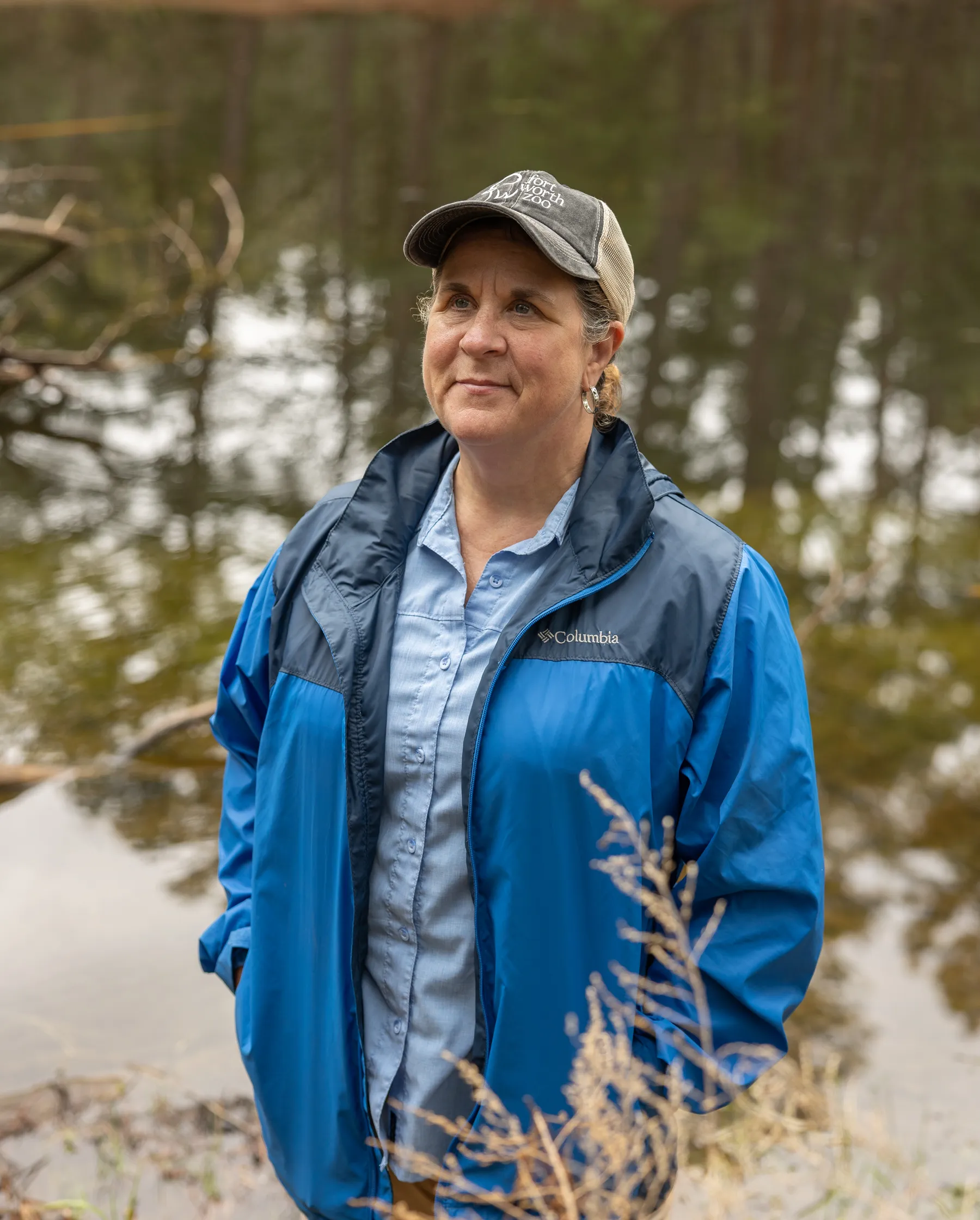 A middle-aged woman wearing a blue jacket and hat stands near a pond