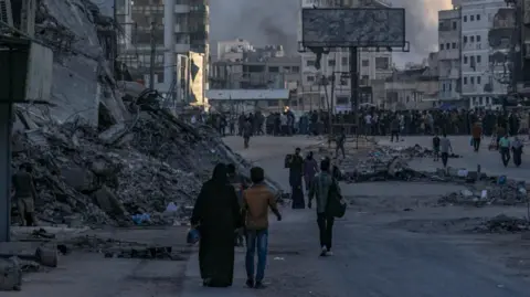 EPA A Palestinian man and woman walk down a street littered with debris and rubble in Gaza City. In the distance, there is a crowd of people, destroyed buildings and smoke billowing into the sky.