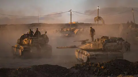 Getty Images Israeli soldiers prepare tanks on August 18, 2025 near the Gaza Strip's northern borders, Israel. Large plumes of dust in orange evening light. 