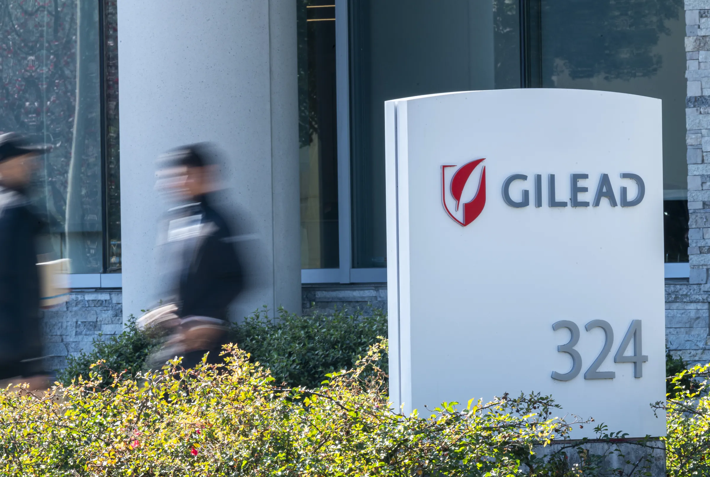 The exterior of Gilead Sciences headquarters in Foster City, California, with people walking past a sign displaying the company’s logo.