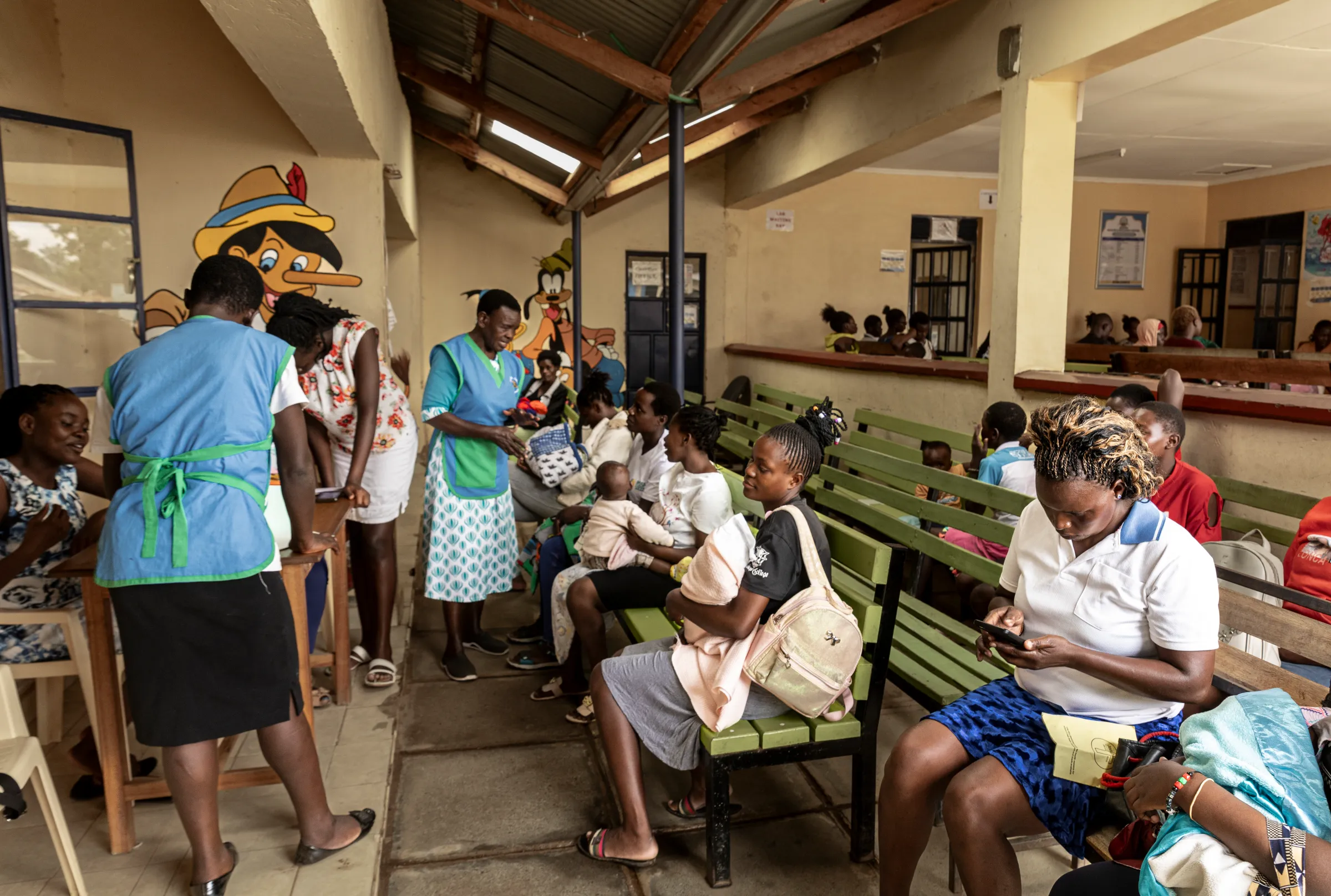 Mothers sit on benches in a busy clinic waiting area in Kisumu, Kenya, while nurses in blue uniforms assist patients at the maternal and child health unit.