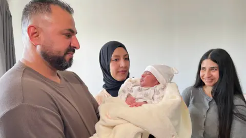 BBC Ahmed with his wife, Nour and teenage daughter, Rahaf. They are all happy and focused on a sleeping newborn baby, being held by Ahmed and wrapped in a cream coloured blanket.   