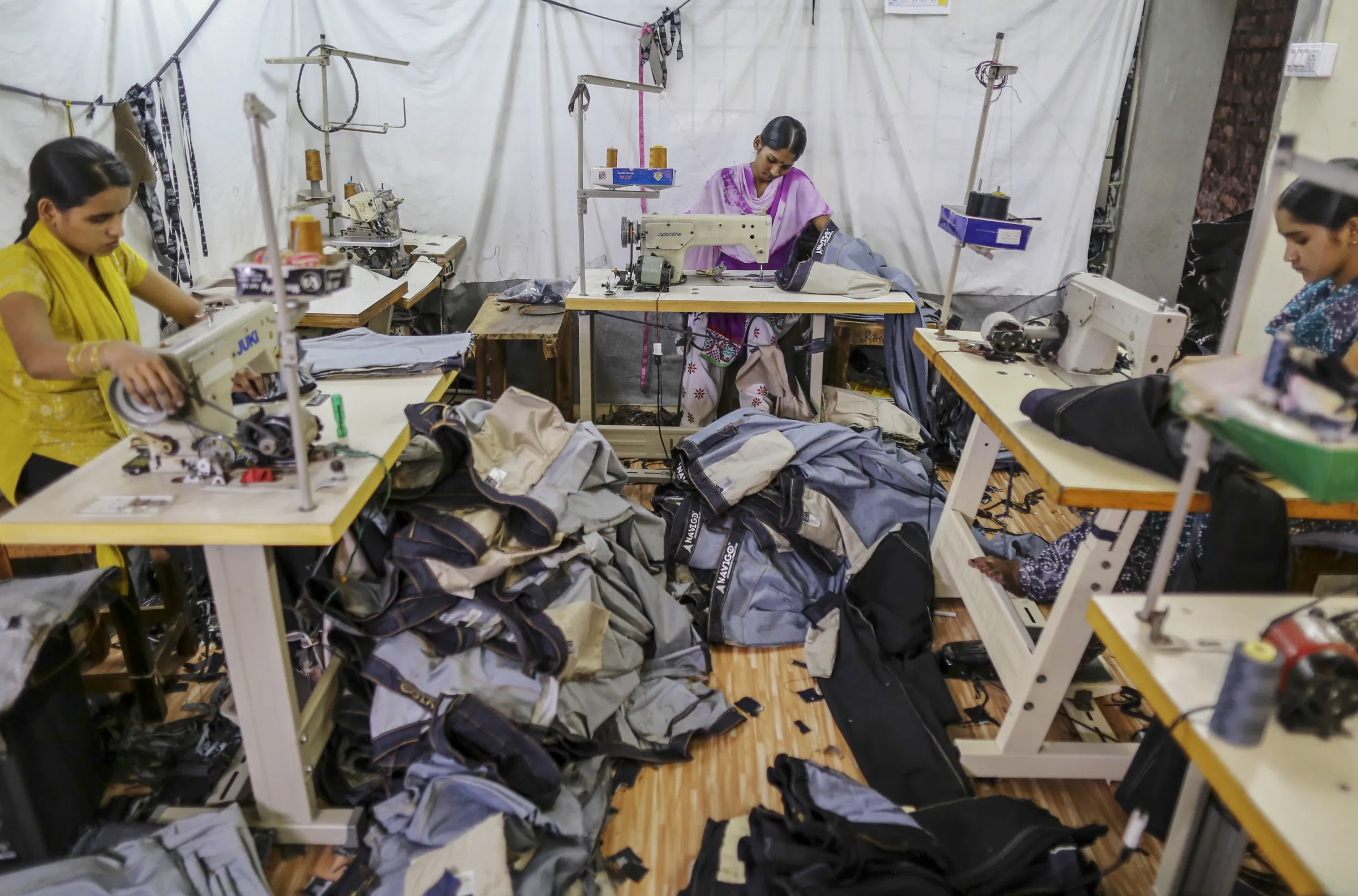 Indian women work on sewing machines in a garment factory, with piles of finished denim clothes in front of them.