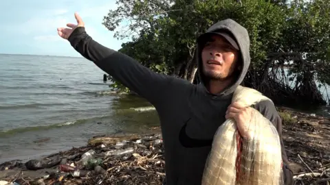 Gustavo Ocando Alex Wilder wears a grey hoodie while standing on a beach littered with bottles and branches. He has pulled the hood over a black baseball cap. Draped over his left shoulder is a fishing net. With his right arm he points towards the sea