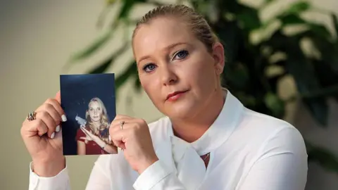 Getty Images Virginia Giuffre, a woman with blonde hair that has been tied back, wearing a white blouse and holding up a photograph of herself as a teenager