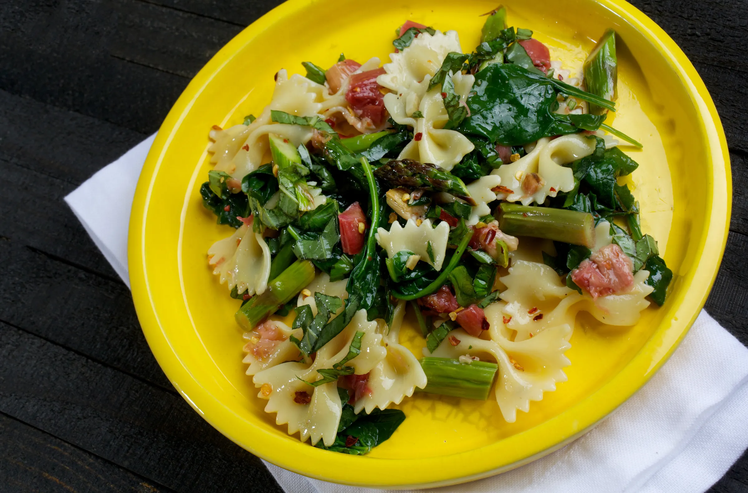 A plate of pasta, bacon, and greens on a yellow plate