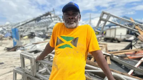 Brandon Drenon / BBC A man wearing a bright yellow shirt with the Jamaican flag on it is seen leaning on metal debris. Everything behind him is destroyed