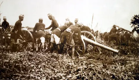 National Army Museum A sepia-toned photo shows men in military gear handling a cannon. 