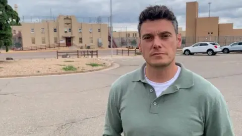 A man in a light green shirt stands holding a microphone outside a light brown brick building surrounded by high fences