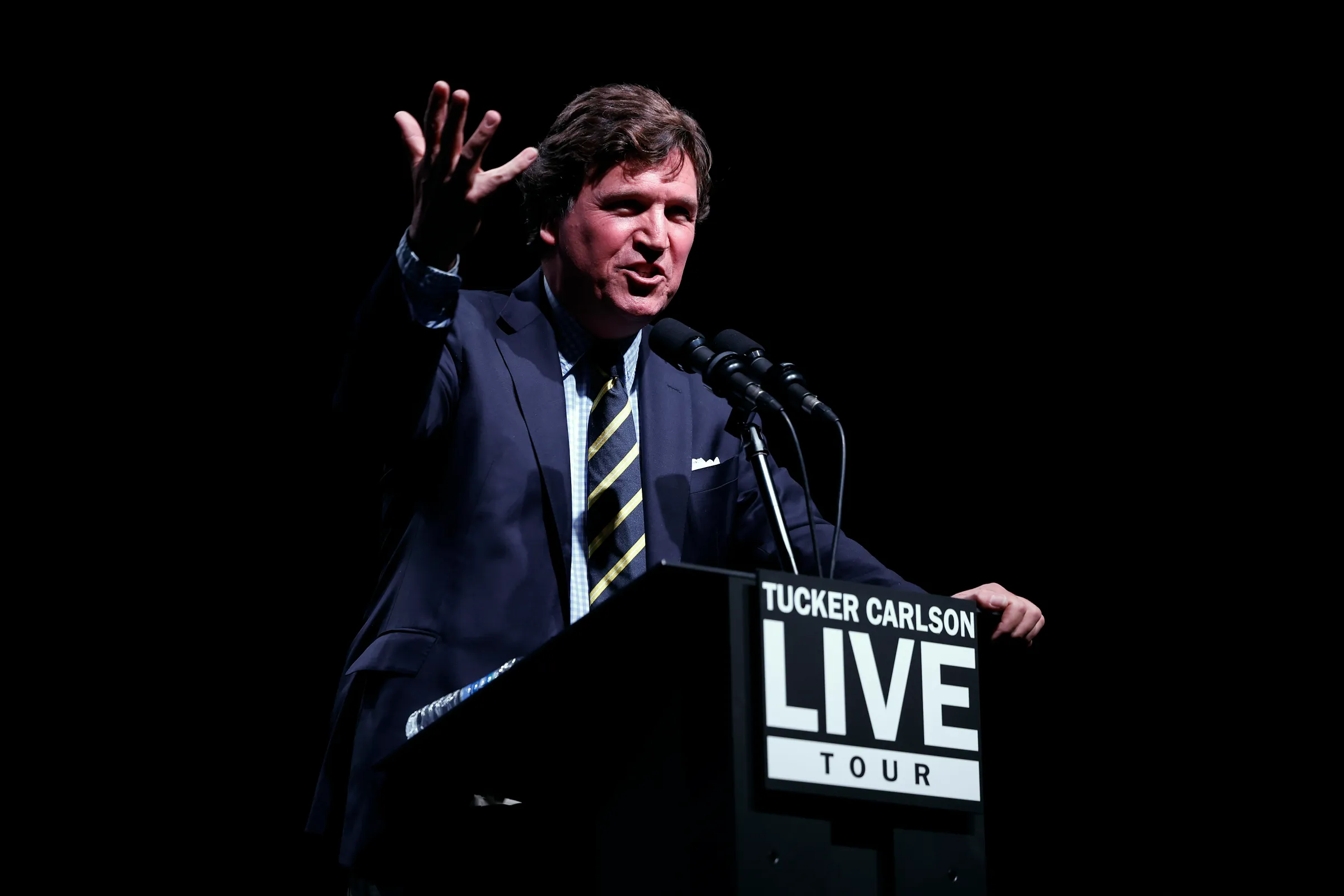 Tucker Carlson, smiling and gesturing, speaks from a podium that reads “Tucker Carlson Live Tour.”