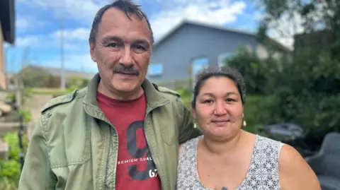 Ulrik and Johanne are pictured outdoors. In the background, are a couple of buildings, some greenery and a path sloping upwards. Ulrik is dressed in a green jacket with a red T-shirt underneath while Johanne is wearing an open-neck patterned dress. 
