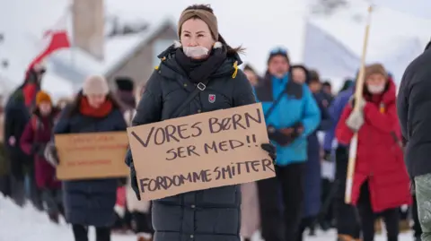 Getty Images Protesters, dressed in thick winter coats with snow in the background, carry placards during a demonstration in Nuuk, Greenland. The protester at the front of the image has tape covering her mouth.