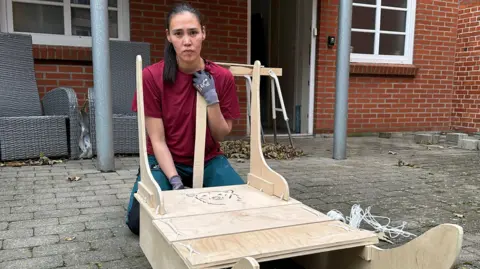 Keira, who is dressed in a red T-shirt and wearing protective gloves, is crouched behind the wooden sleigh she's been making. In the background are a couple of grey garden chairs and an open door leading into a home.
