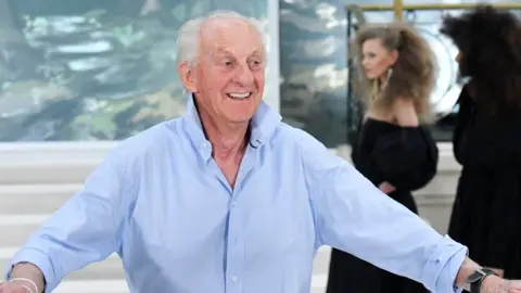 Getty Images Paul Costelloe is is smiling broadly and has receding grey hair. He is wearing a blue open-neck shirt and his arms are stretched out on either side. behind him are three female models dressed in black.