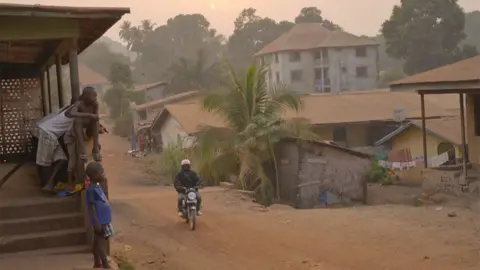 A motorbike travelling along a dirt road in Sierra Leone with children looking on from a porch