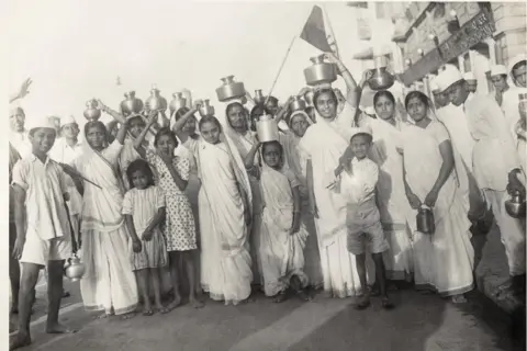 Alkazi Collection of Photography The monochrome photo shows a group of children, many of whom are carrying steel pots filled with sewater on their heads. The boys are dressed in loose shirts, shorts and caps, while the girls are wearing saris. 
