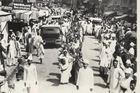 The Alkazi Collection of Photography A wide-angle shot of India's bustling streets as men and women, most of whom are protesters, walk through the traffic along a boulevard dotted with shops