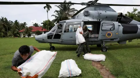 Reuters A young man carries a sack of aid from a helicopter across a green field.
