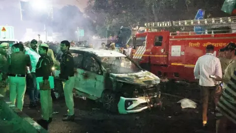 RAJAT GUPTA/EPA/Shutterstock Indian police personnel inspect the scene of a car explosion. A white burnt out car can be seen, while a red fire engine is in the background