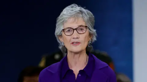 Reuters Catherine Connolly, with short grey hair, wearing a purple top and purple jacket. She is standing behind a wooden podium. 