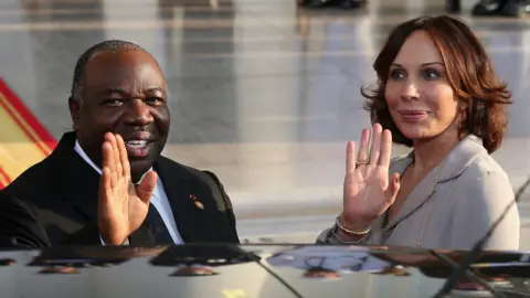 Getty Images Gabon President Ali Bongo Ondimba in a black suit and spouse Sylvia Bongo Ondimba in a grey suit, both waving