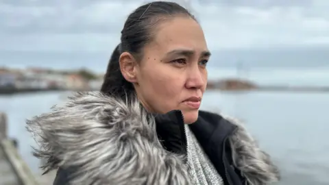 BBC Keira, who has long, dark hair in a ponytail and is wearing a coat with a furry collar, stares into the distance. Behind her is a body of water, probably the sea. 