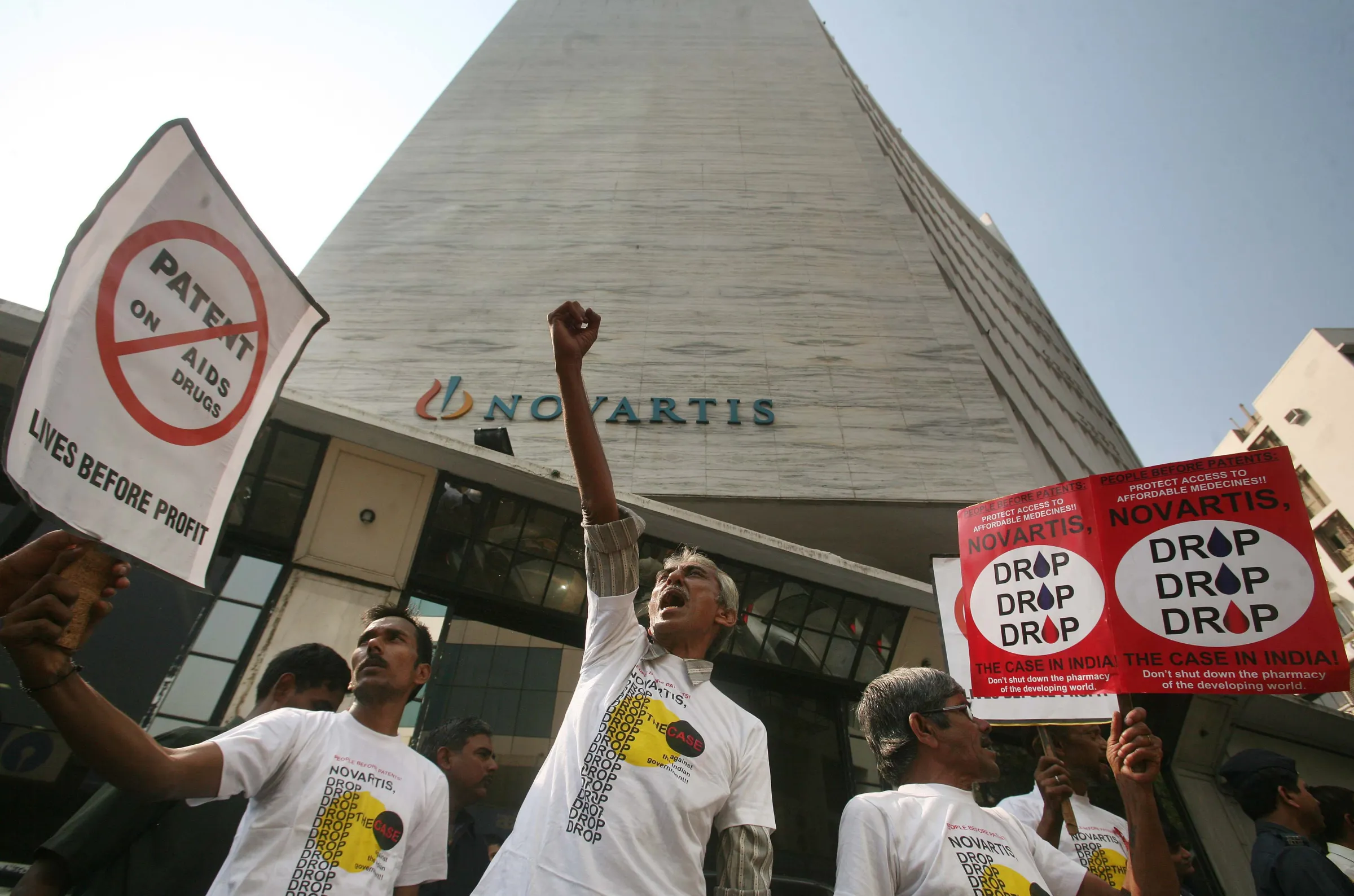 HIV-rights activists stand in front of a tall Novartis office building in Mumbai, holding signs that read “No patents on AIDS drugs” and “Drop the case,” during a street protest.
