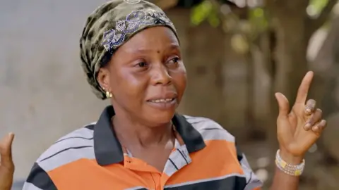 BBC Sally Kalokoh in a headscarf and shirt with white, black and orange stripes and a black collar gestures with her hands. On one of her arms she wears a beaded pearl-like bracelet.