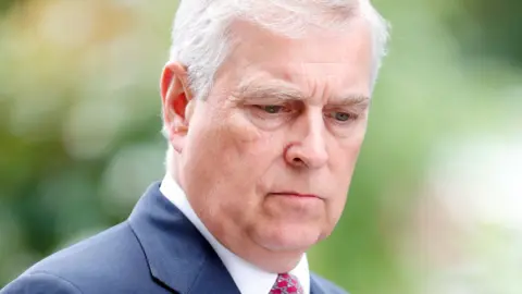 Indigo / Getty Images A close head and shoulders crop shows Andrew looking down towards the floor while wearing a suit and red tie against a neutral green background with heavy bokeh, in Ascot in 2019.