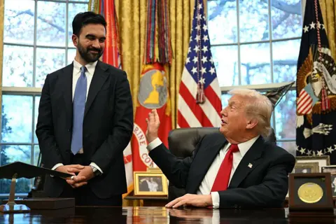 Getty Images Mamdani is seen standing next to Trump, who is seated behind his desk in the Oval Office. Both are smiling and Trump is reaching out to jokingly tap him on the arm