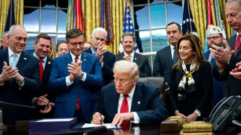 EPA US President Donald Trump signs document to end shutdown, holding it up at his desk which a crowd of people around him applaud.