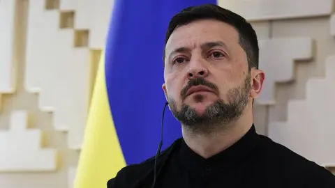 Getty Images Ukraine's president Volodymyr Zelensky looks out during a press conference. His face is stern. He is wearing a black jacket and shirt. The Ukrainian flag - in blue and yellow - is displayed on the wall behind him.