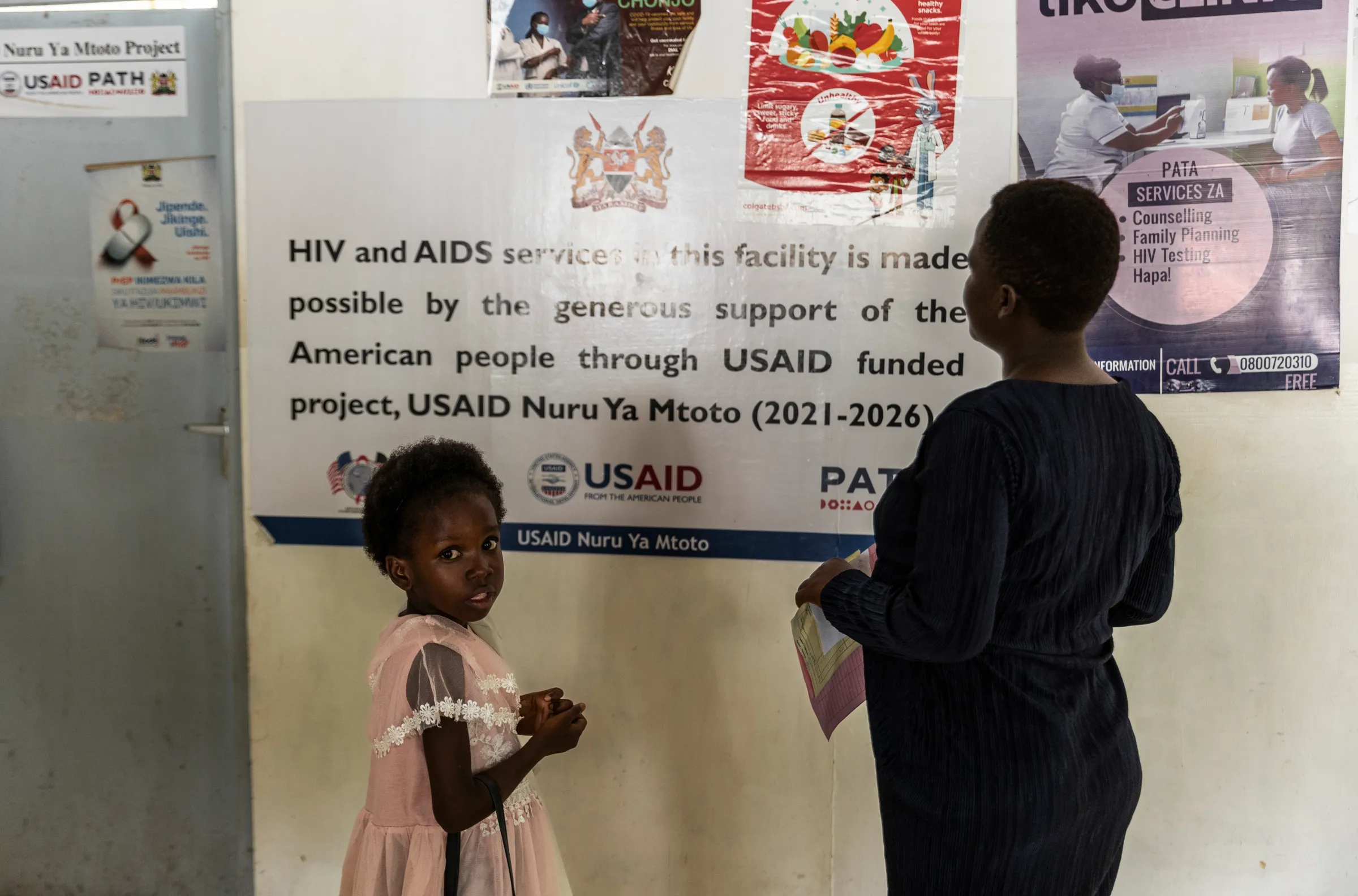 A mother and daughter stand in front of a sign at a USAID-funded health facility.