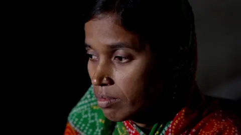 Seraj Ali Lalmunni Devi pictured wearing a multi-coloured saree and sitting in a dark room. She lost her husband to toxic alcohol consumption in 2022. 