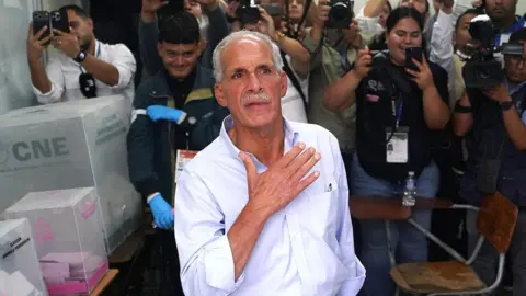 AFP via Getty Images An image showing Tito Asfura at a polling station in Honduras. Behind him are spectators who have their phones out to capture the moment. Asfura is in the centre of the image, wearing a white button-up shirt rolled at the sleeves. He is gesturing with his right hand towards his chest. 
