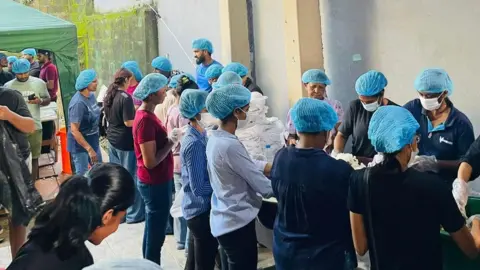 Sasindu Sahan Tharaka Rows of people, all wearing blue hairnets, working in a makeshift kitchen