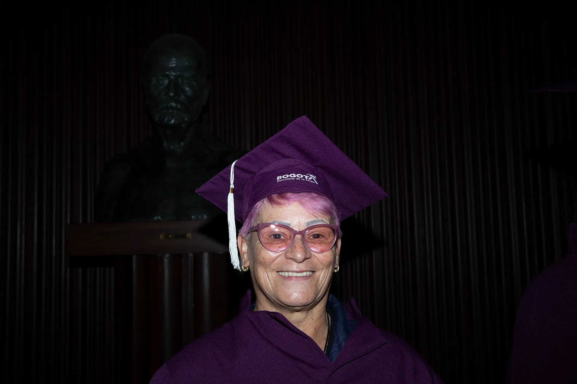 a woman with pink hair and pink glasses smiles while wearing a purple graduation cap and gown