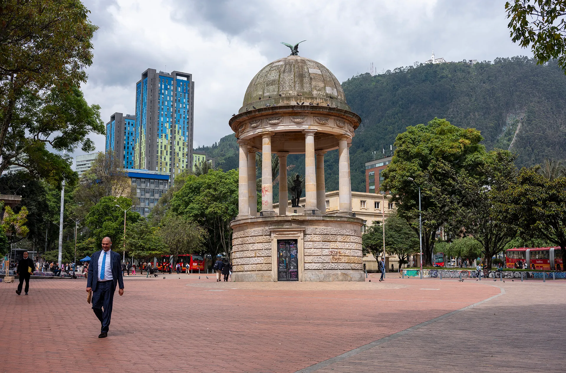 a gilded statue of Simón Bolívar in a Bogotá square with commuters walking past. Mountains and foliage surrounds
